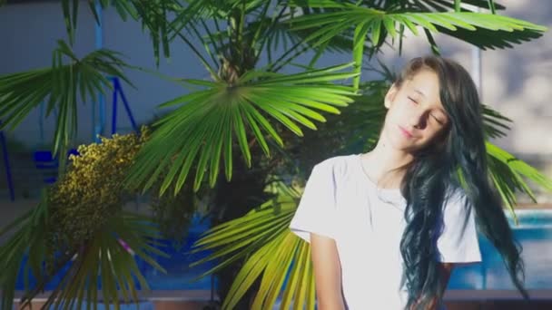 Красивая брюнетка позирует на фоне пальмовых листьев. Девочка-подросток с голубыми длинными кудрявыми волосами. летний солнечный день. 4k, slow motion — стоковое видео