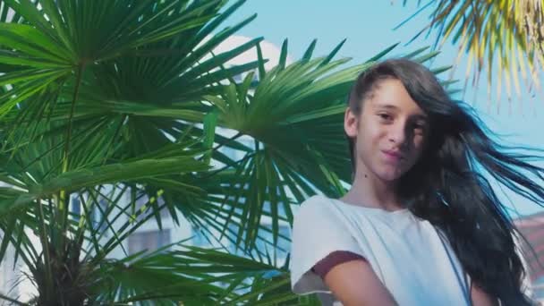Vackra brunett flicka poserar mot bakgrund av palm lämnar. En flicka Tonåring med blå långt lockigt hår. solig sommardag. 4k, Slowmotion — Stockvideo