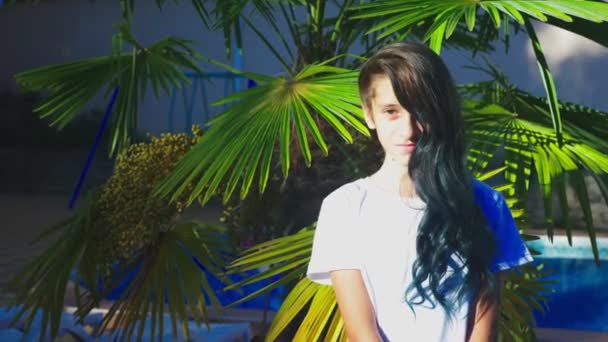 Krásná bruneta dívka pózuje na pozadí palm listy. Dospívající dívka s modrými dlouhé kudrnaté vlasy. letní slunečný den. 4k, pomalý pohyb — Stock video