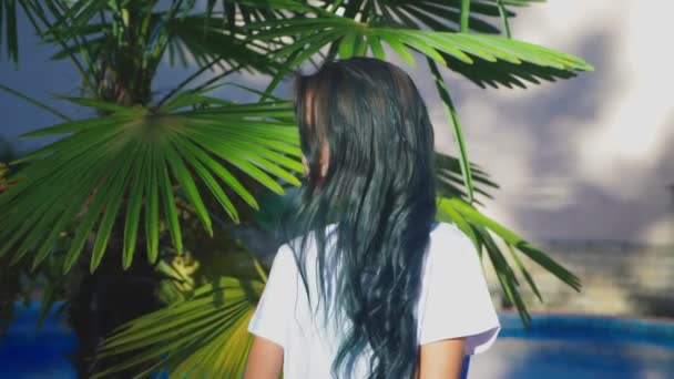 Piękna brunetka dziewczyna pozowanie na tle palm liści. Nastolatek dziewczyna z niebieski długie kręcone włosy. słoneczny letni dzień. 4k, zwolnionym tempie — Wideo stockowe