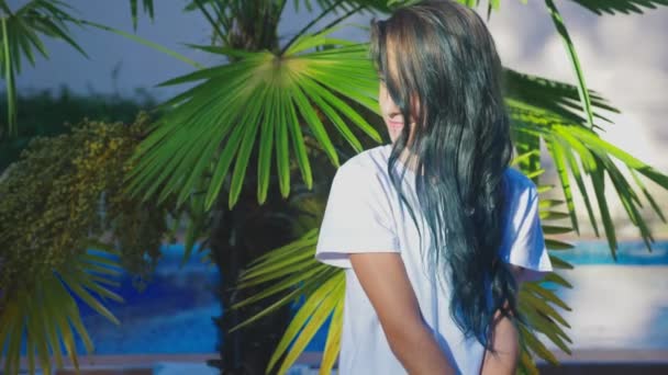 Schöne brünette Mädchen posiert vor einem Hintergrund von Palmblättern. ein Mädchen mit blauen langen lockigen Haaren. Sommersonniger Tag. 4k, Zeitlupe — Stockvideo