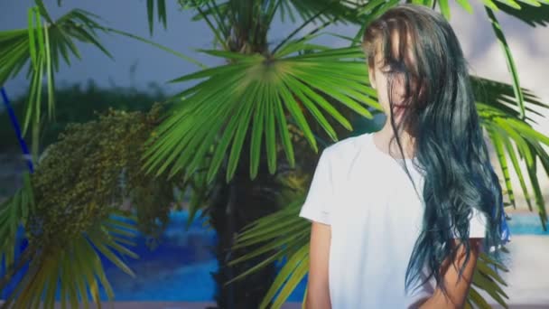 Красива брюнетка дівчина позує на тлі пальмового листя. Дівчинка-підліток з синім довгим кучерявим волоссям. літній сонячний день. 4k, повільний рух — стокове відео