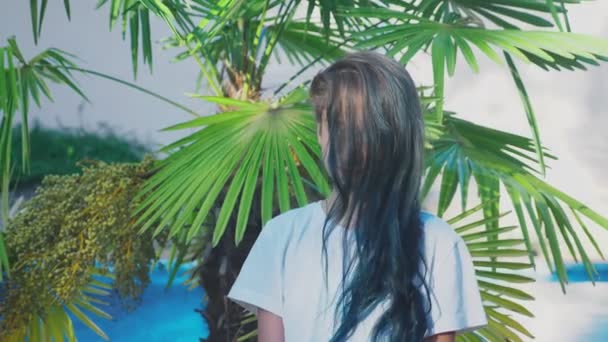 Mooie brunette meisje poseren tegen een achtergrond van palm bladeren. Tiener meisje met blauwe lang krullend haar. zonnige zomerdag. 4k, slow-motion — Stockvideo