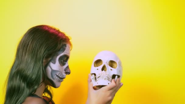 Halloween, fille avec squelette maquillé sur la moitié du visage, habillée en sorcière, posant sur un fond jaune vif. 4k, ralenti, gros plan — Video