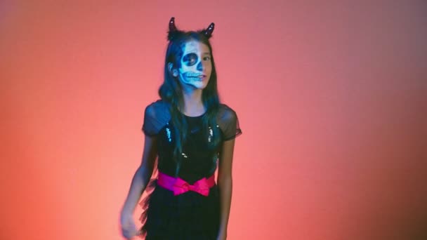 Хеллоуїн, дівчина з макіяжним скелетом на половині обличчя, одягнена як відьма, позує і танцює на червоному тлі. 4k, повільний рух — стокове відео