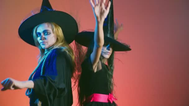 Halloween, en flicka med smink, ett skelett för halva ansiktet och hennes mor, klädd som häxor, poserar och dansar på en röd bakgrund. 4k, Slowmotion — Stockvideo
