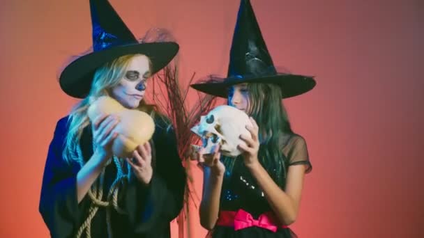 Halloween, uma menina com maquiagem, um esqueleto para metade do rosto e sua mãe, vestida como bruxas, posando e dançando em um fundo vermelho. 4k, câmera lenta — Vídeo de Stock