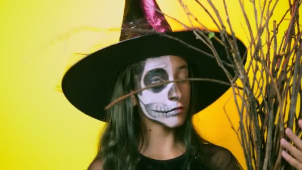 Halloween, fille avec squelette maquillé sur la moitié du visage, habillée en sorcière, posant sur un fond jaune vif. 4k, ralenti, gros plan — Video