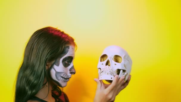 Halloween, chica con esqueleto de maquillaje en media cara, vestida de bruja, posando sobre un fondo amarillo brillante. 4k, cámara lenta, primer plano — Vídeo de stock