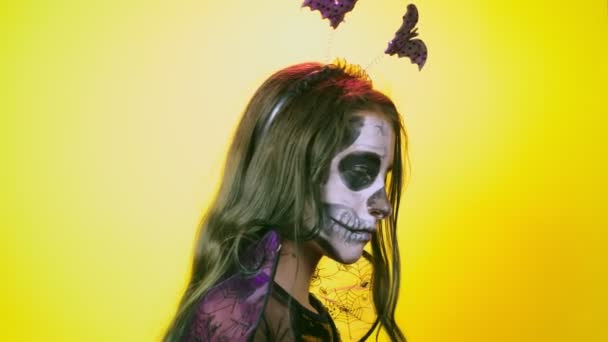 Хеллоуїн, дівчина з макіяжним скелетом на половині обличчя, одягнена як відьма, позує на яскраво-жовтому тлі. 4k, повільний рух, крупним планом — стокове відео