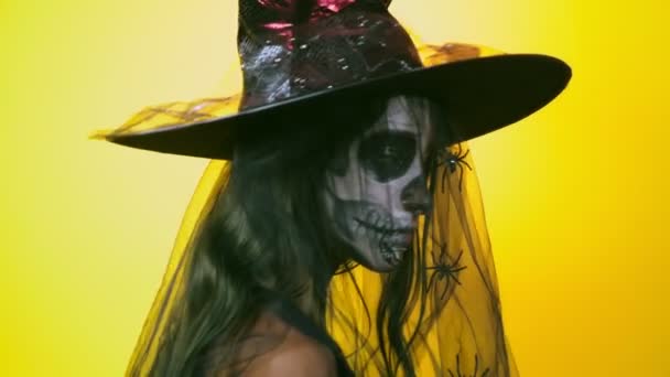 Halloween, girl with make-up skeleton on half face, dressed as witch, posing on bright yellow background. 4k, slow-motion, close-up — Stock Video