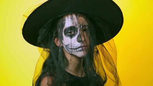 Halloween, fille avec squelette maquillé sur la moitié du visage, habillée en sorcière, posant sur un fond jaune vif. 4k, ralenti, gros plan — Video