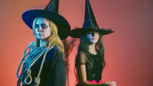 Хэллоуин, девушка с макияжем, скелет для половины лица и ее мать, одетые как ведьмы, позирующие и танцующие на красном фоне. 4k, slow motion — стоковое видео