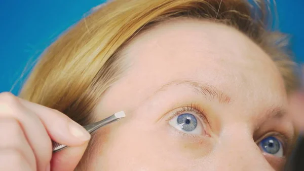 Extremo close-up. arrancando pinças de sobrancelhas, uma menina loira com olhos azuis puxa cabelos desnecessários de sua sobrancelha — Fotografia de Stock