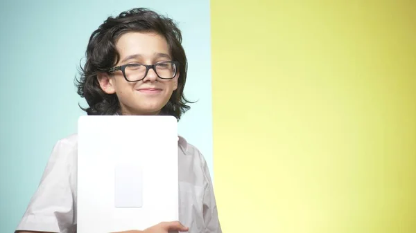 Ritratti di un adolescente in uniforme scolastica e occhiali su sfondo colorato. Che tipo divertente. concetto di apprendimento. Un adolescente è in possesso di un computer portatile, guardando la fotocamera, sorridendo e facendo facce divertenti — Foto Stock