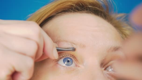 Primer plano extremo. arrancando las pinzas de las cejas, una chica rubia con ojos azules saca pelos innecesarios de su ceja, cámara lenta . — Vídeos de Stock