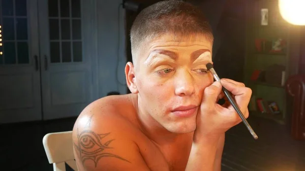 Concepto de maquillaje parodia. un joven guapo se pone maquillaje en la cara, sentado frente a un espejo en el vestidor. artista travesti profesional. primer plano , — Foto de Stock