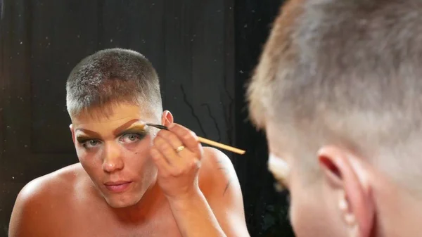 Koncepcja parodia makijażu. przystojny, młody człowiek stosuje makijażu na twarzy, siedząc przed lustrem w szatni. parodia profesjonalnego artysty. szczelnie-do góry, — Zdjęcie stockowe