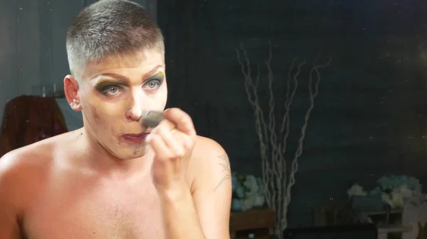 Concepto de maquillaje parodia. un joven guapo se pone maquillaje en la cara, sentado frente a un espejo en el vestidor. artista travesti profesional. primer plano , — Foto de Stock