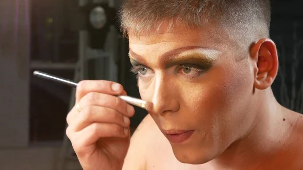 Concepto de maquillaje parodia. un joven guapo se pone maquillaje en la cara, sentado frente a un espejo en el vestidor. artista travesti profesional. primer plano , —  Fotos de Stock