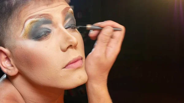 Begreppet parodi make-up. en stilig ung man applicerar makeup i ansiktet, sitter framför en spegel i omklädningsrummet. professionell travesti konstnär. närbild, — Stockfoto