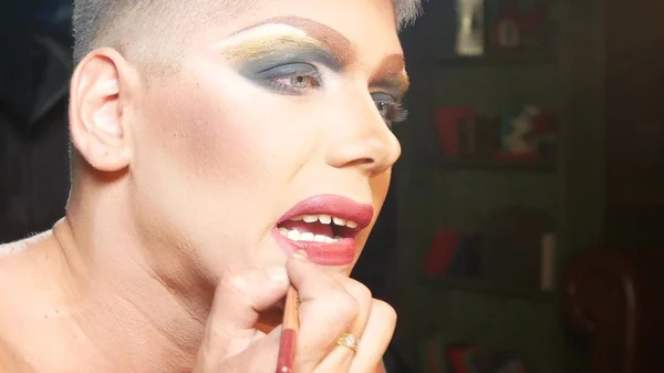 Concepto de maquillaje parodia. un joven guapo se pone maquillaje en la cara, sentado frente a un espejo en el vestidor. artista travesti profesional. primer plano , — Foto de Stock