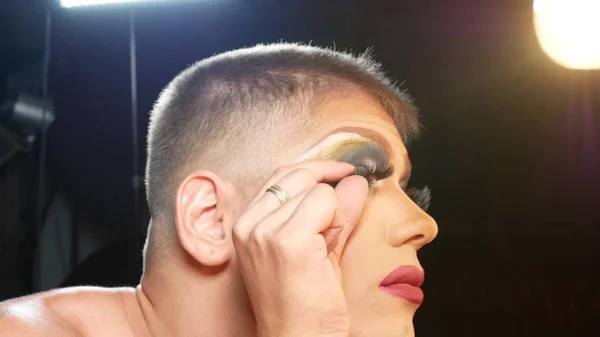 Concept of travesty make-up. a handsome young man applies makeup on his face, sitting in front of a mirror in the dressing room. professional travesty artist. close-up, — Stock Photo, Image