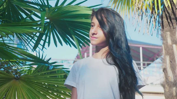 Piękna brunetka dziewczyna pozowanie na tle palm liści. Nastolatek dziewczyna z niebieski długie kręcone włosy. słoneczny dzień lata. — Zdjęcie stockowe