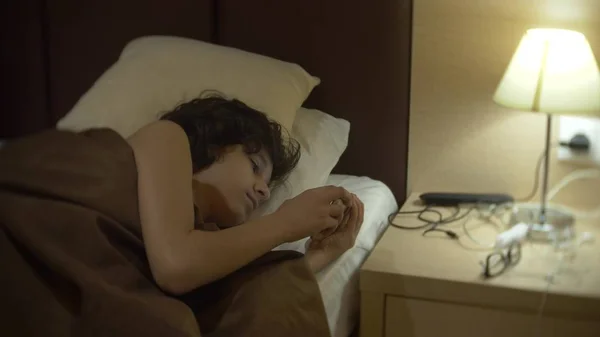 Schattige tiener jongen in zijn kamer 's nachts, liggend op het bed met behulp van een smartphone. zijn nachtlampje brandt — Stockfoto