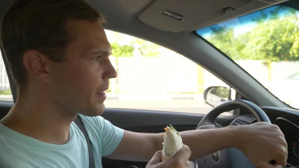 El hombre al volante del coche, come comida rápida, mientras el coche se mueve. en un día soleado de verano . — Foto de Stock