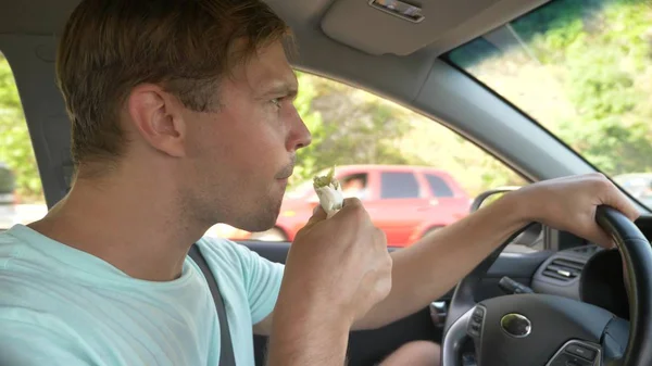 El hombre al volante del coche, come comida rápida, mientras el coche se mueve. en un día soleado de verano . — Foto de Stock