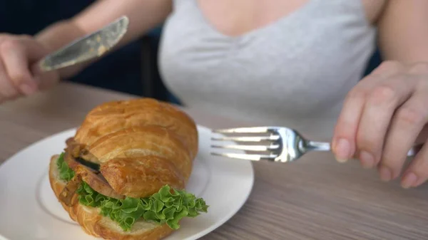 Eine unkenntliche Frau mit schönem Dekolleté frühstückt in einem Café mit einem Croissant-Sandwich mit gekochtem Schweinefleisch und trinkt Kaffee. Nahaufnahme. — Stockfoto