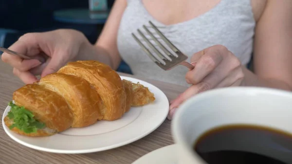 An unrecognizable woman with a beautiful neckline, breakfasts in a cafe with a croissant sandwich with boiled pork and drinking coffee. close-up. — Stock Photo, Image