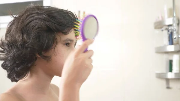 Söt pojke borsta hans lockiga hår framför badrumsspegeln. — Stockfoto