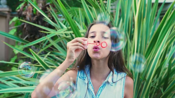 Chica adolescente morena soplando jabón burbujas contra un tropical parque fondo . —  Fotos de Stock