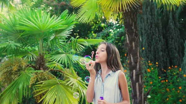 Gadis remaja berambut cokelat meniup gelembung sabun terhadap latar belakang taman tropis . — Stok Foto