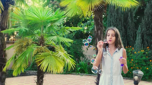 Chica adolescente morena soplando jabón burbujas contra un tropical parque fondo . —  Fotos de Stock