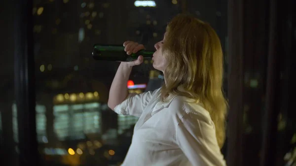 Una mujer está bebiendo cerveza en casa junto a una ventana panorámica con vistas a los rascacielos. noche, fondo borroso . — Foto de Stock