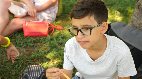 Attraktiver Junge isst einen frischen Burger, der auf Gras unter einem Baum im Park sitzt. — Stockfoto