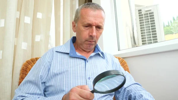 Un hombre un pensionista lee un libro a través de una lupa en casa junto a la ventana . — Foto de Stock