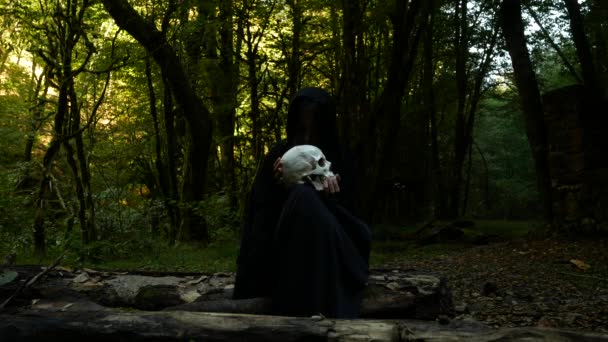 Straszne zła czarownica w lesie jesienią trzyma ludzką czaszkę, siedząc na dziennik. — Wideo stockowe