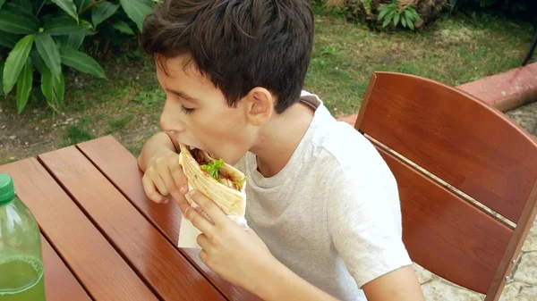 Um menino atraente come um pita de pão fresco com uma salada de vegetais frescos e carne, sentado em um café fast food sob uma árvore no parque. . — Fotografia de Stock