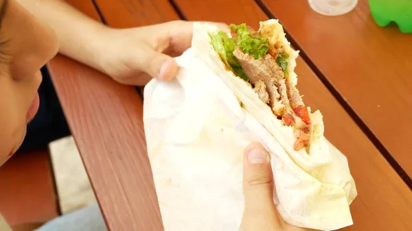 Um menino atraente come um pita de pão fresco com uma salada de vegetais frescos e carne, sentado em um café fast food sob uma árvore no parque. . — Fotografia de Stock