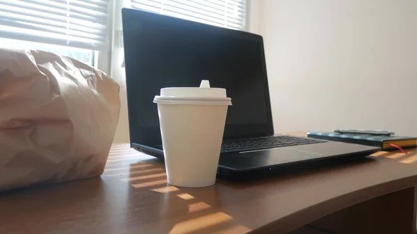 Un escritorio de hombres de negocios con una taza desechable para café y comida rápida. ordenador portátil abierto. desayuno en la oficina, los rayos del sol hacen su camino a través de las persianas, espacio de copia — Foto de Stock