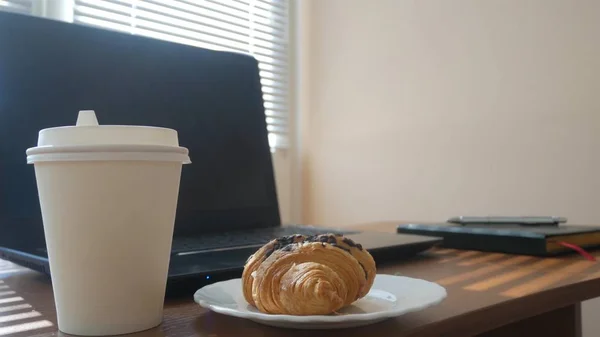 Un bureau d'affaires avec une tasse jetable pour le café et la restauration rapide. ordinateur portable ouvert. petit déjeuner au bureau, les rayons du soleil font leur chemin à travers les stores, copier l'espace — Photo