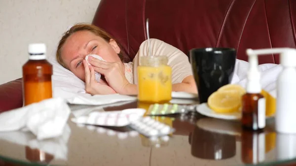 Nemocná žena leží na pohovce ve svém obývacím pokoji, na stole, vedle ní jsou léky a horký nápoj s medem a citronem. — Stock fotografie