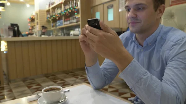 Joven guapo usando un teléfono inteligente, bebiendo café y sonriendo mientras se relaja en un café. Tiempo de Navidad — Foto de Stock