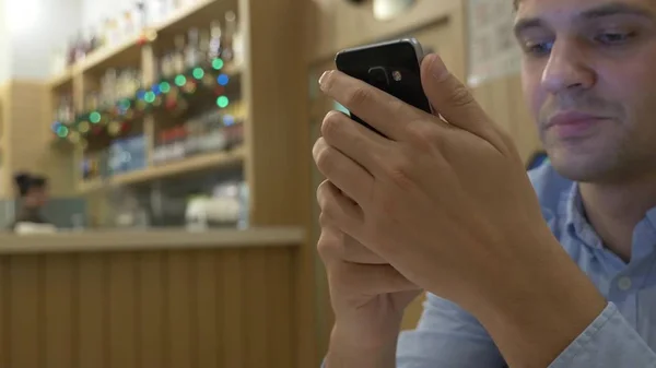 Joven guapo usando un teléfono inteligente, bebiendo café y sonriendo mientras se relaja en un café. Tiempo de Navidad — Foto de Stock