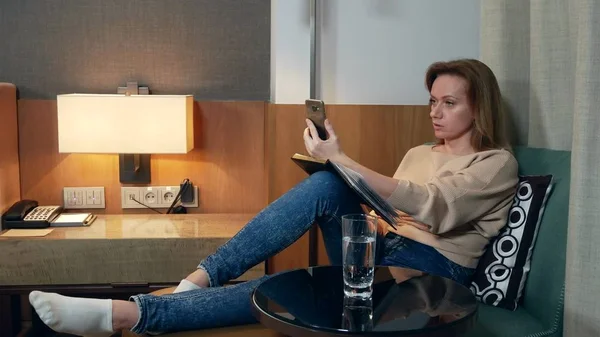Ввечері жінка сидить на зручному кріслі у вітальні, розмовляє на відео зі свого смартфона і робить нотатки в блокноті — стокове фото