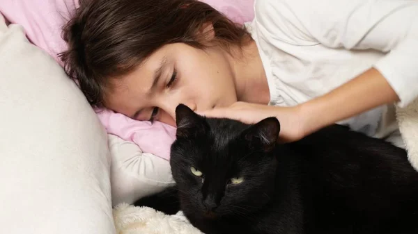 Una niña pequeña abraza y acaricia a su mascota, un gato negro, acostado en la cama — Foto de Stock